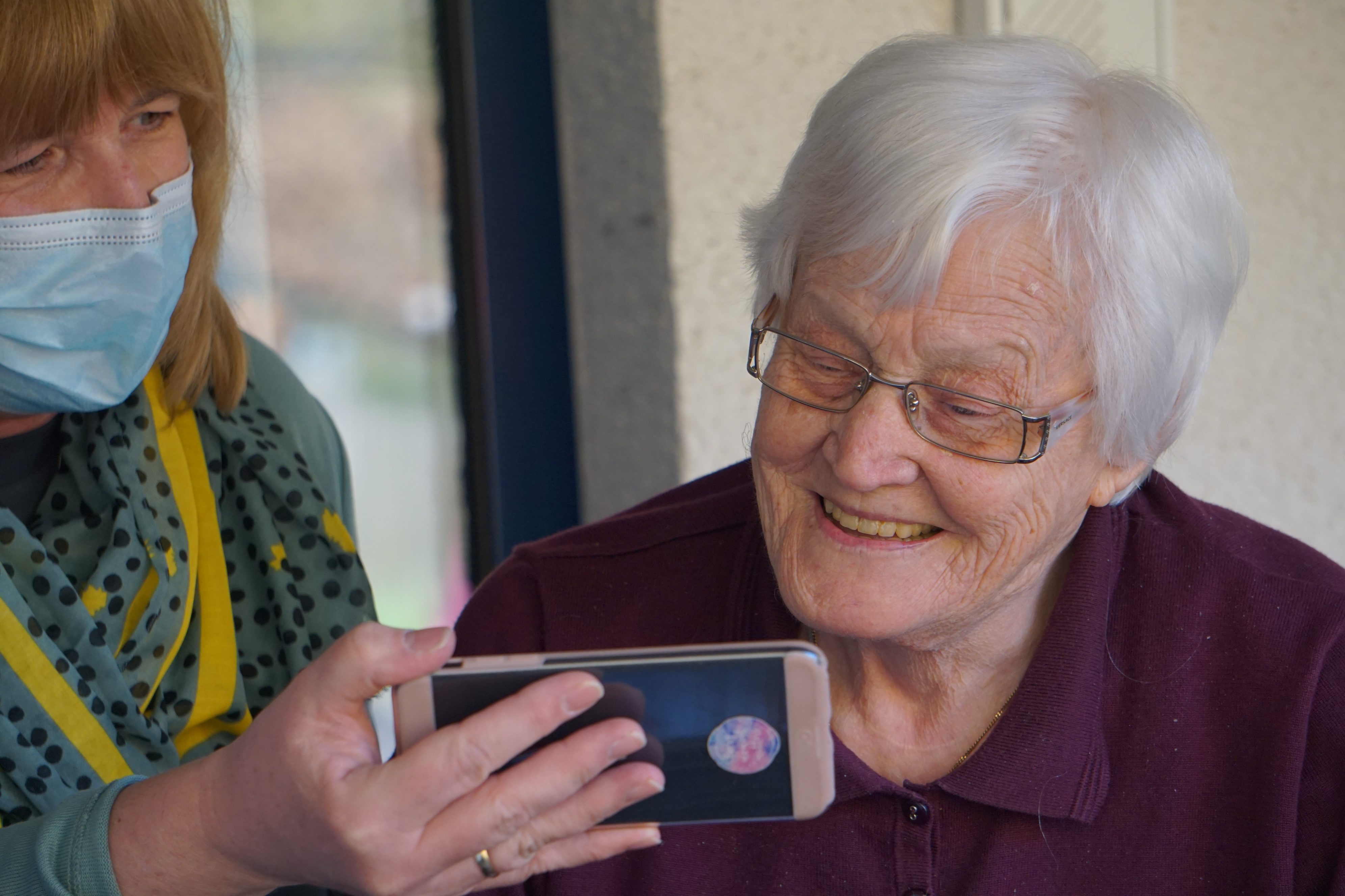 Narrative Medicine Care image showing a care Supporter holding a smartphone up for an elderly woman to see as they talk.